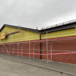 Elewacja budynku Biedronki z żółtą ścianą i czerwoną cegłą, osłonięta rusztowaniem z taśmą ostrzegawczą, przygotowana do prac remontowych.