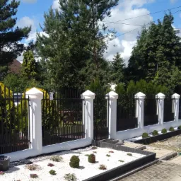 Nowoczesne ogrodzenie z czarnymi przęsłami i białymi słupkami, rabata z białym grysem i młodymi roślinami, widok z poziomu chodnika, fragment zaparkowanego czerwonego samochodu w tle.