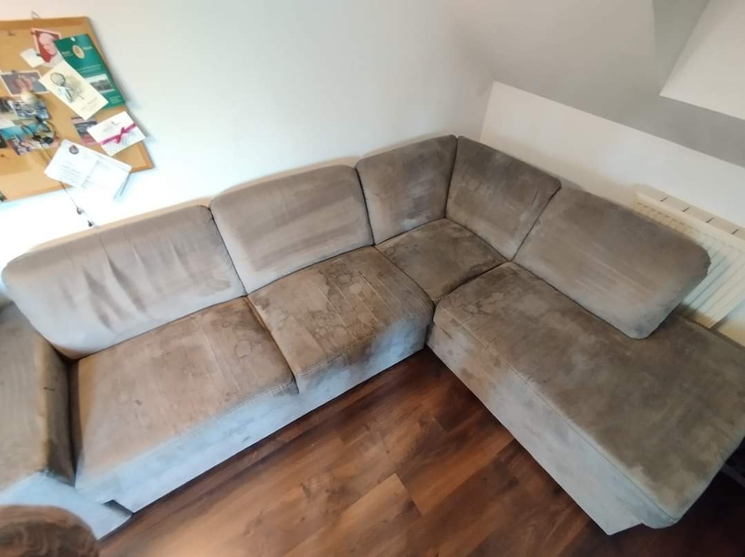 Narożnik z szarej tapicerki z widocznymi plamami przed czyszczeniem, na drewnianej podłodze, widoczna tablica korkowa na ścianie.