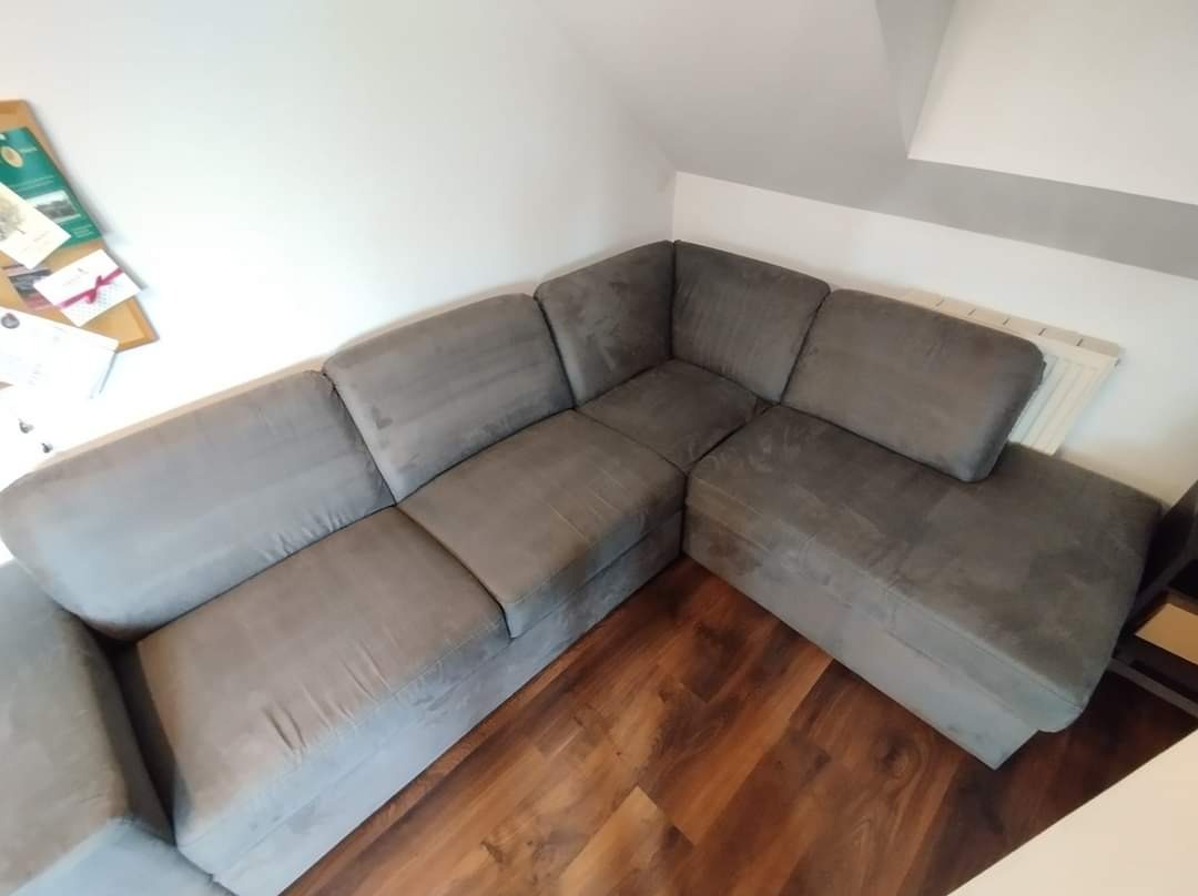 Szara, narożna kanapa z widocznymi śladami czyszczenia na jednym z segmentów, ukazująca różnicę w kolorze i strukturze materiału, umieszczona w rogu pokoju z drewnianą podłogą.