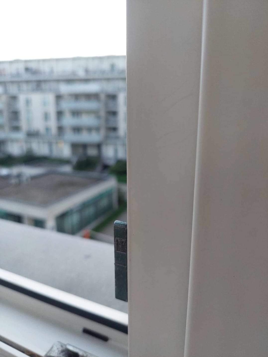 Biała rama okienna z widocznymi zarysowaniami i brudnym parapetem, z zewnątrz widok na budynki mieszkalne w tle.