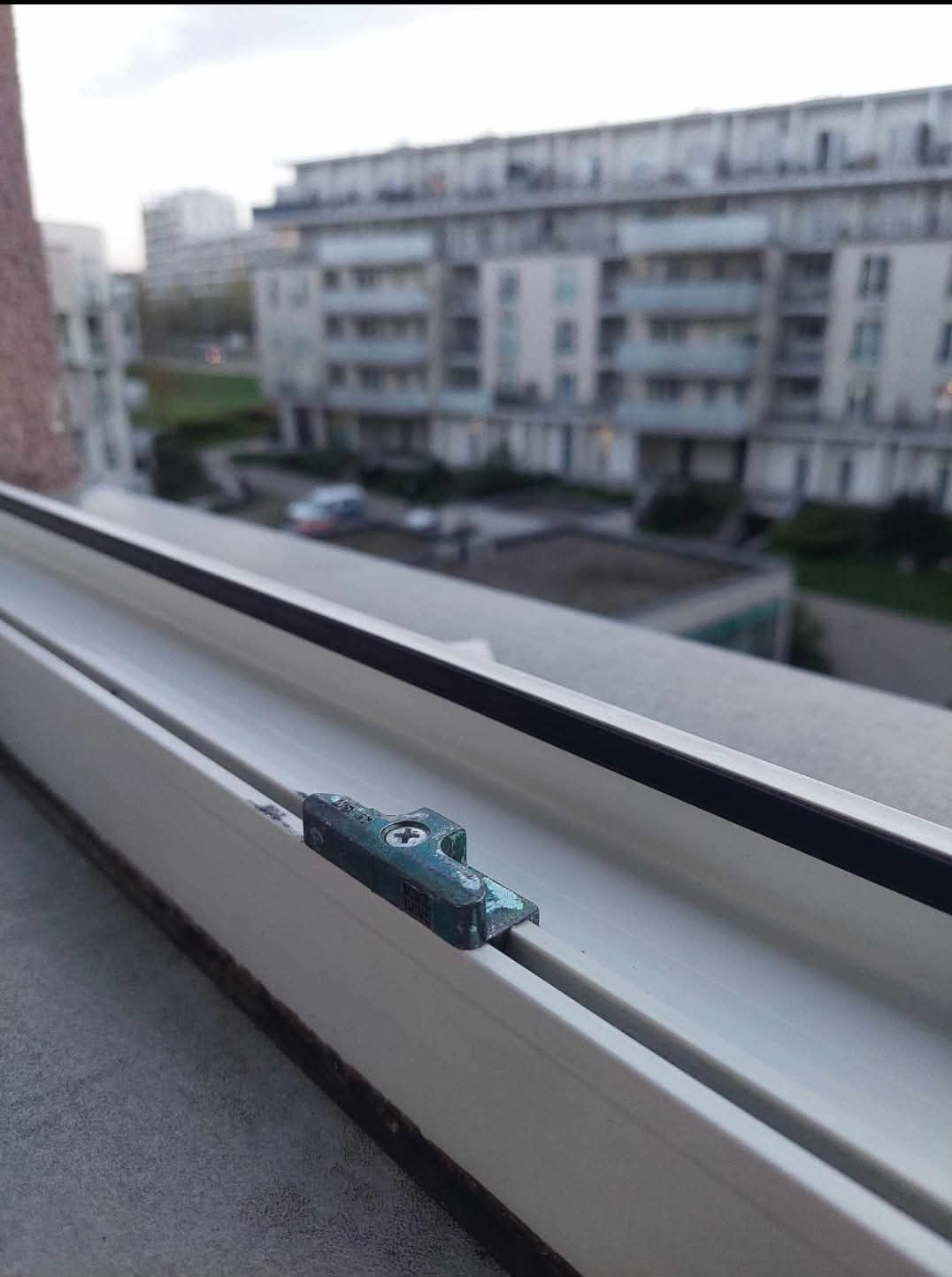 Detail zardzewiałego okucia okiennego w oknie PCV, widok na osiedle mieszkalne w tle.