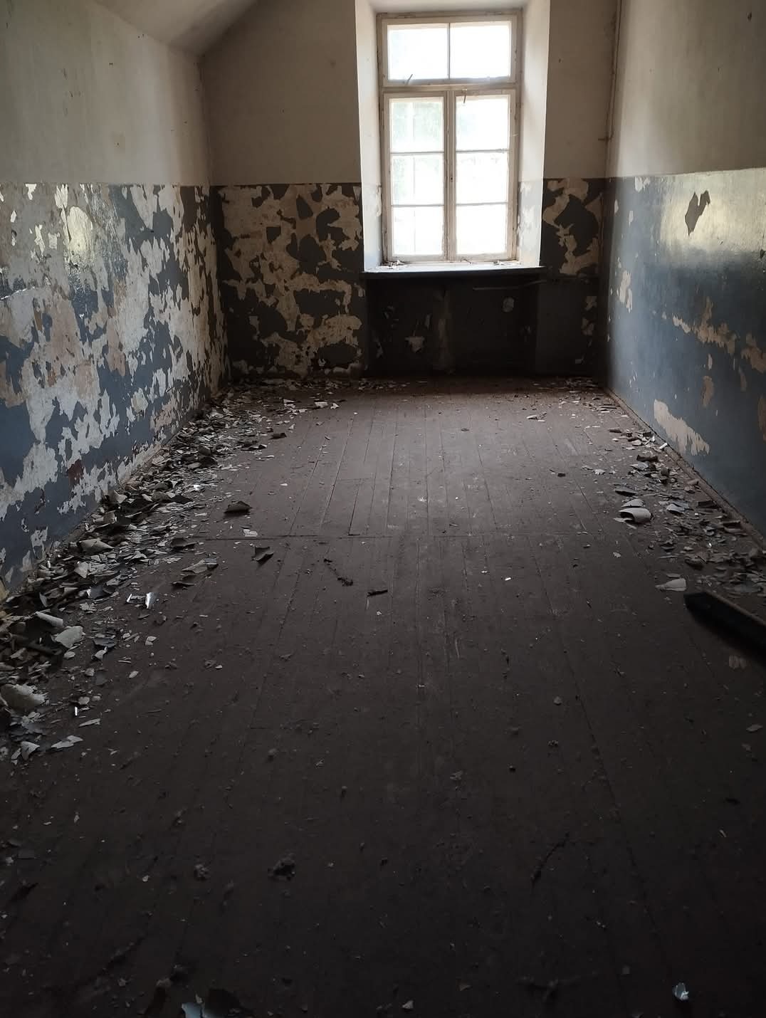 Pusty pokój z odpadającą farbą na ścianach i zabrudzoną podłogą z desek, widok na okno z drewnianą ramą.