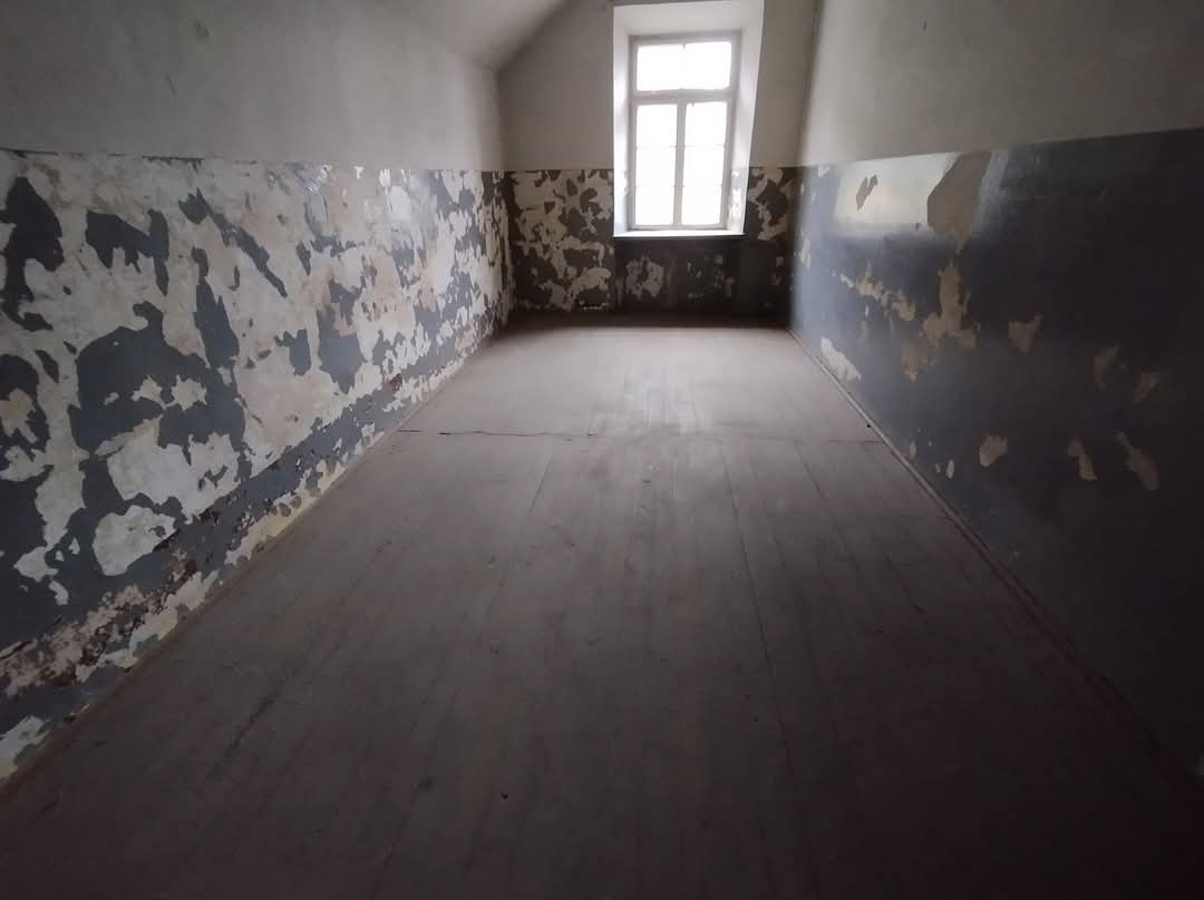 Puste pomieszczenie z łuszczącą się farbą na ścianach i drewnianą podłogą, widoczne ślady po remoncie, małe okno w tle.