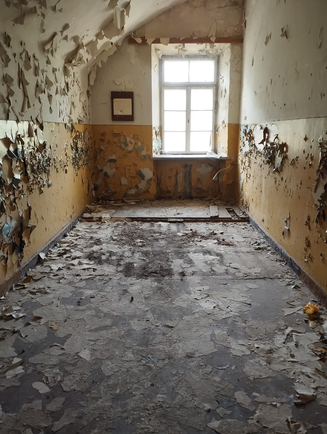 Pokój na poddaszu w stanie dewastacji: odpadająca farba, resztki na podłodze, małe okno w głębi.