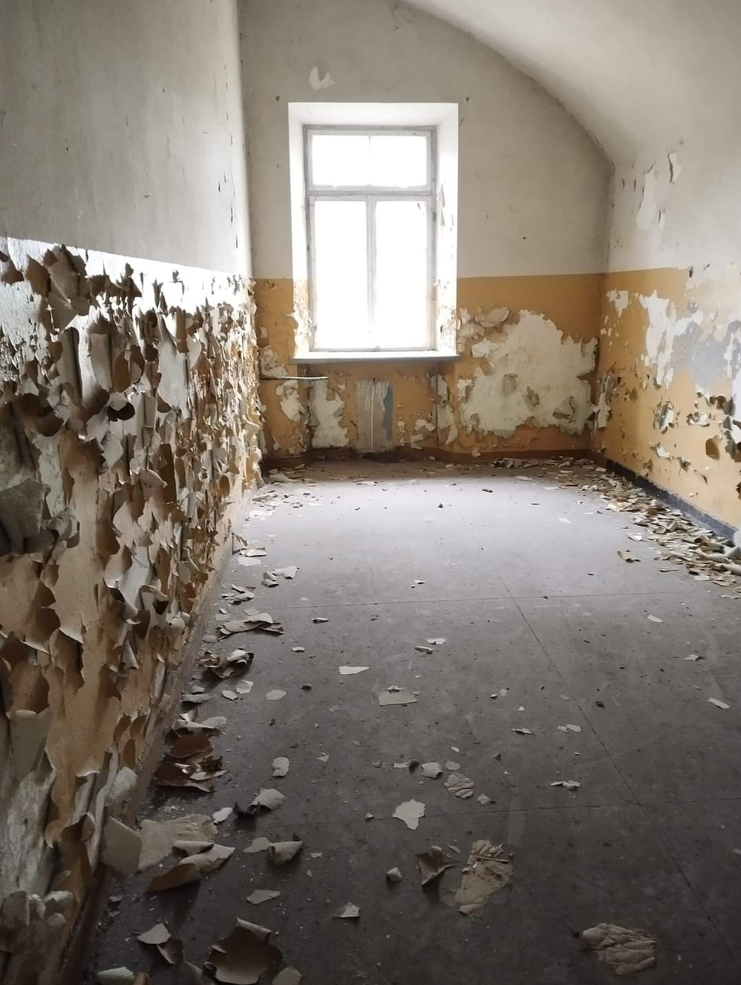 Pokój w opuszczonym budynku z odpadającą farbą od ściany, widoczne fragmenty farby na podłodze, okno w tle.
