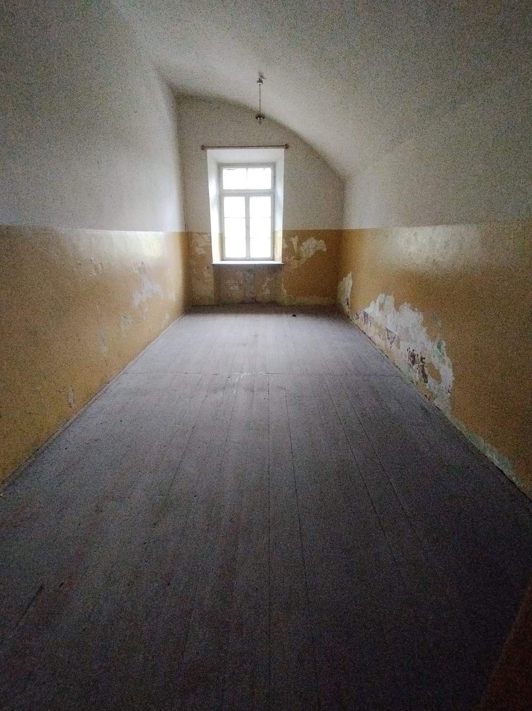 Pusty strych w Warszawie przed remontem. Widoczne zniszczone ściany z resztkami farby, drewniana podłoga i okno w szczycie dachu.