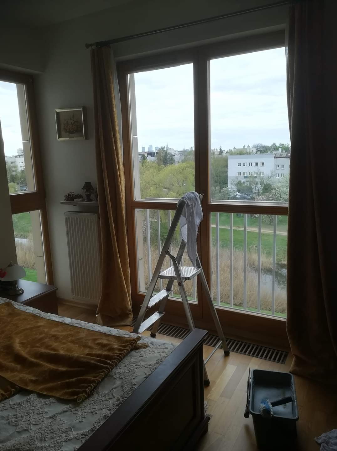 Drabina oparta o okno z widokiem na park i budynki miejskie, obok łóżko z narzutą i wiaderko z narzędziami do czyszczenia.