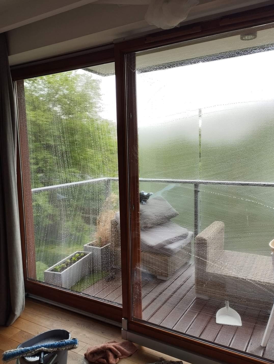 Mycie okien balkonowych z widokiem na zieleń, widoczne smugi wody na szybach, narzędzia do mycia okien w rogu pomieszczenia.