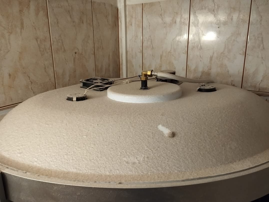 Gruba warstwa pyłu pokrywa okrągły zbiornik w pomieszczeniu z kafelkami, widoczne przewody i elementy instalacji na powierzchni.