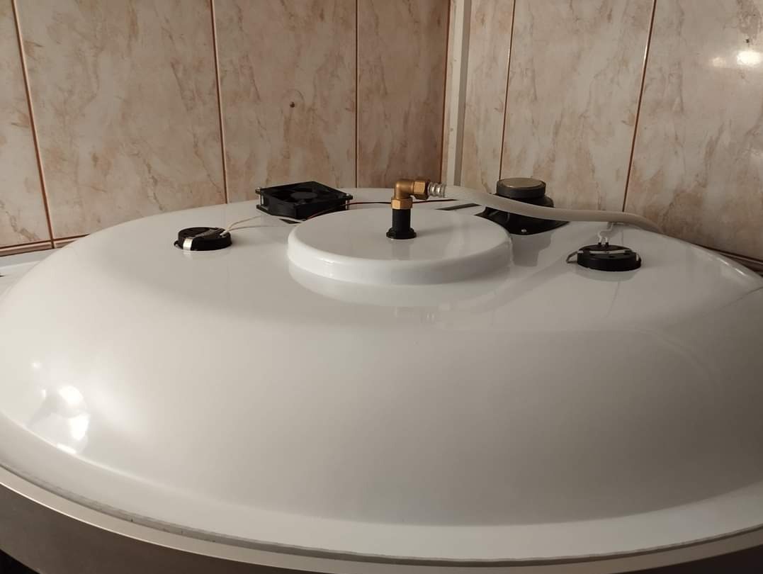 Biały zbiornik z elementami instalacji, wentylatorem i pokrywami na tle płytek ceramicznych.