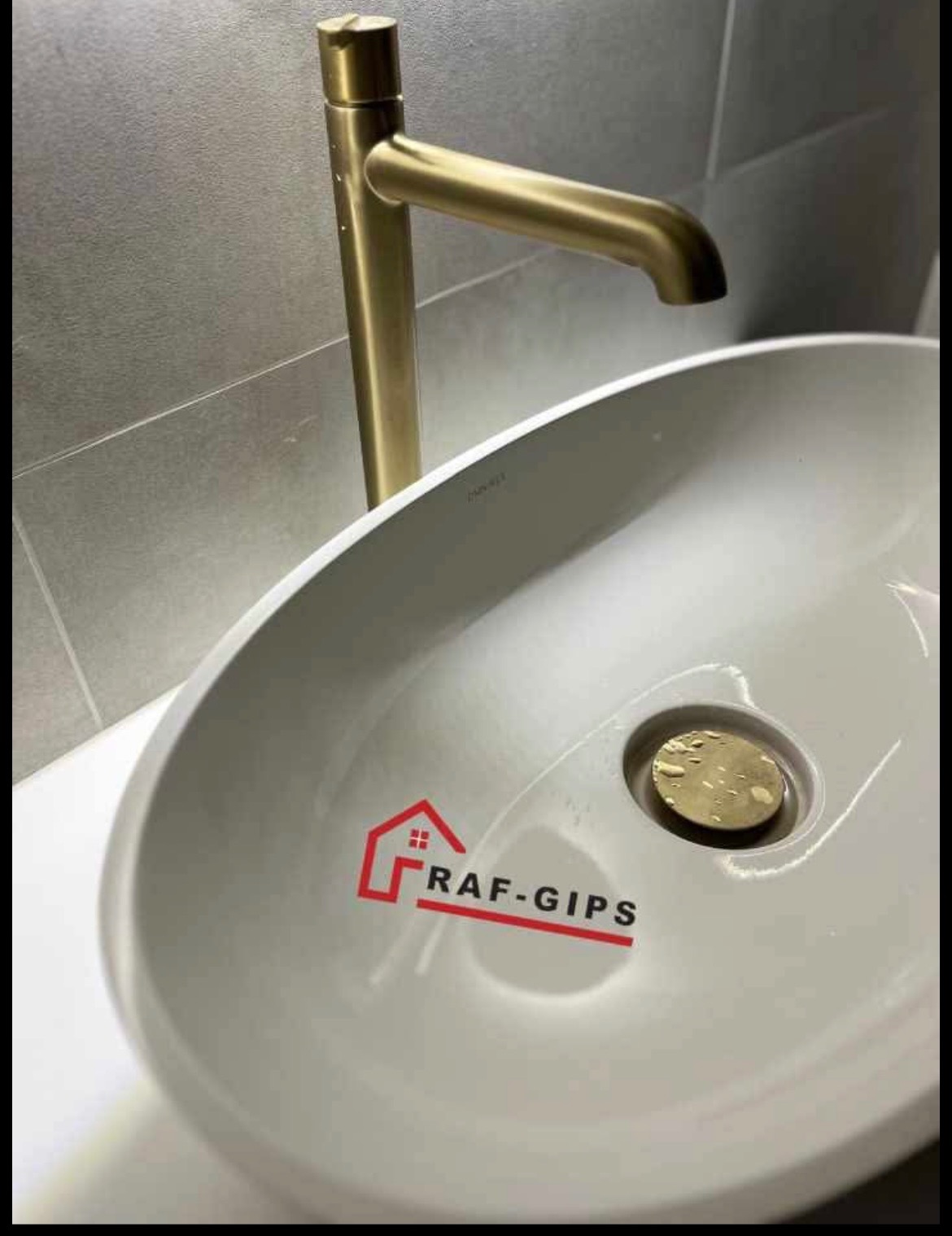 Umywalka ceramiczna z baterią w kolorze szczotkowanego złota, na umywalce naklejka reklamowa firmy RAF-GIPS, szare płytki w tle.