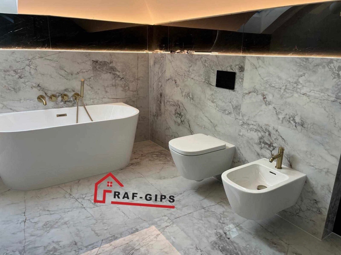 Nowoczesna łazienka z marmurowymi płytkami, wolnostojącą wanną, podwieszaną toaletą i bidetem z złotą armaturą, podświetlany sufit, widoczne logo firmy RAF-GIPS.