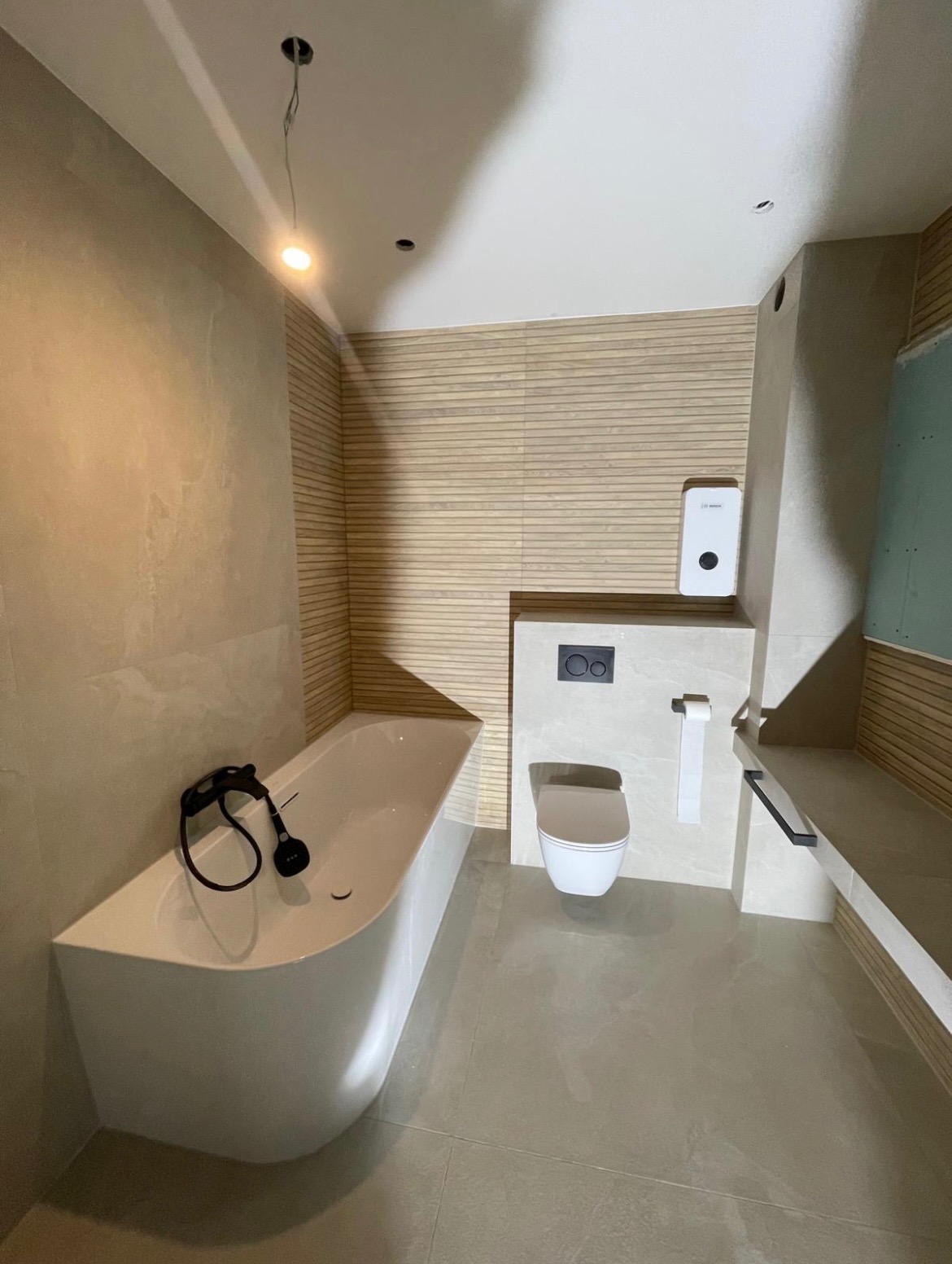 Nowoczesna łazienka z wanną o geometrycznym kształcie, podwieszanym WC, płytkami imitującymi drewno na ścianie i szarą podłogą. Na wannie leżą czarne słuchawki. Widoczne niedokończone wykończenie...