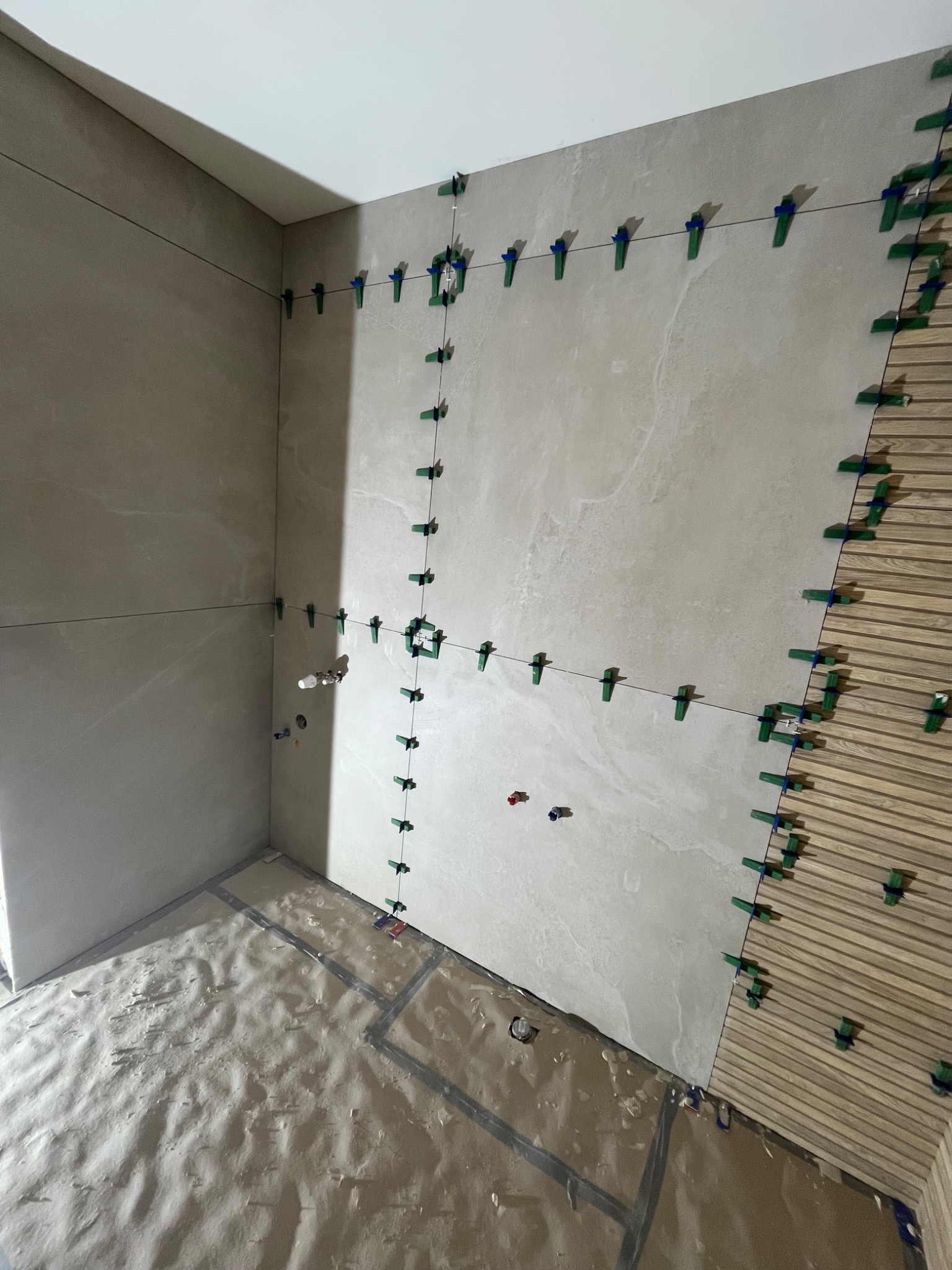 Wnętrze łazienki w trakcie układania płytek ceramicznych i drewnopodobnych paneli ściennych; widoczne systemy poziomowania płytek, sznurki traserskie i zabezpieczona folią podłoga.