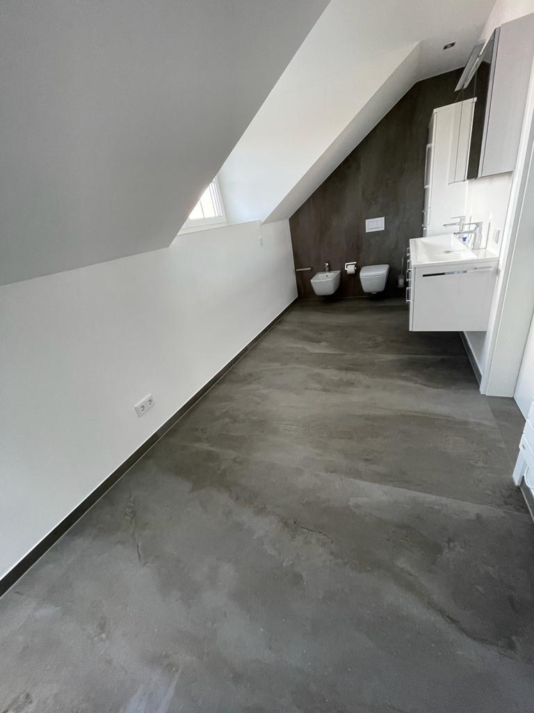 Nowoczesna łazienka na poddaszu z betonową posadzką, białą ceramiką sanitarną i minimalistyczną zabudową umywalkową.