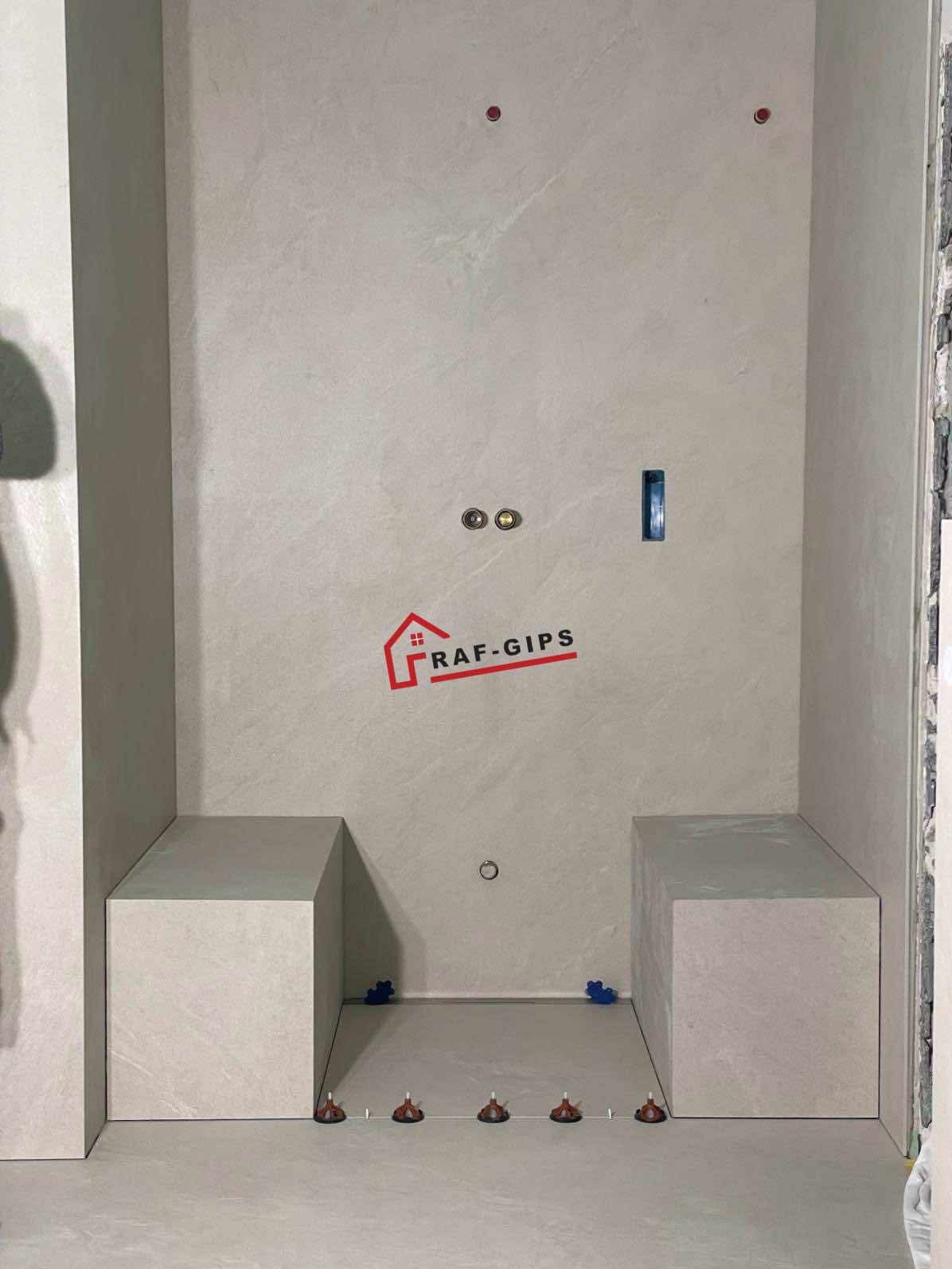 Wnęka prysznicowa w trakcie wykańczania, z ułożonymi płytkami na podłodze i siedziskach, widoczne poziomujące klipsy i logo firmy RAF-GIPS na ścianie.
