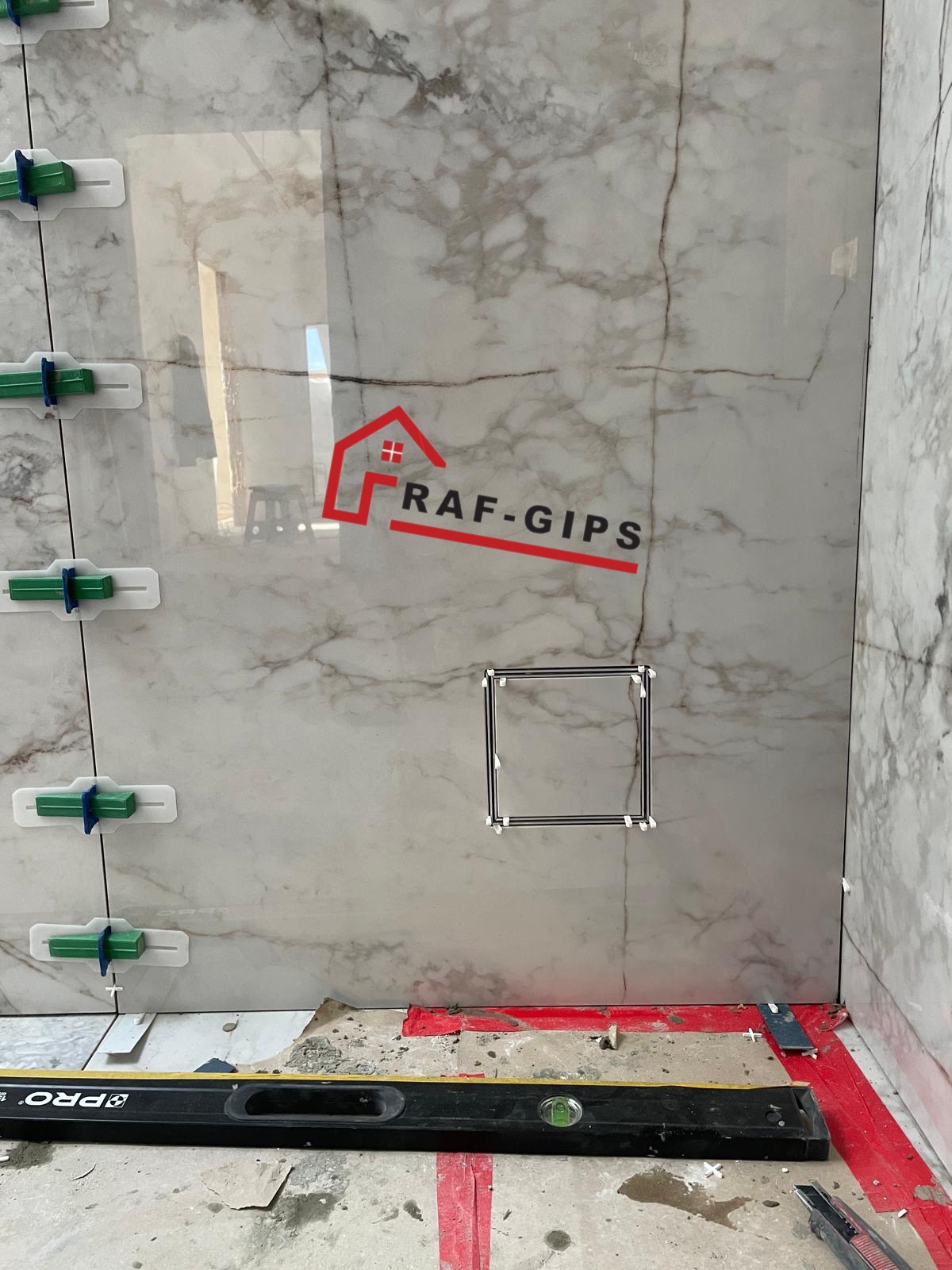 Montaż płytek imitujących marmur w łazience, widoczne klipsy poziomujące, poziomica i logo firmy RAF-GIPS na ścianie.