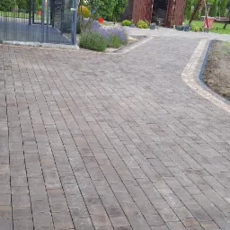 Ułożona kostka brukowa w odcieniach brązu i beżu tworząca ścieżkę prowadzącą od budynku w stronę ogrodu z widoczną przyczepką samochodową.