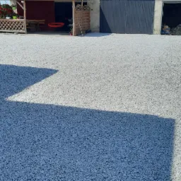 Utwardzona droga żwirowa z ubijarką w tle, widoczne budynki gospodarcze i fragment domu.