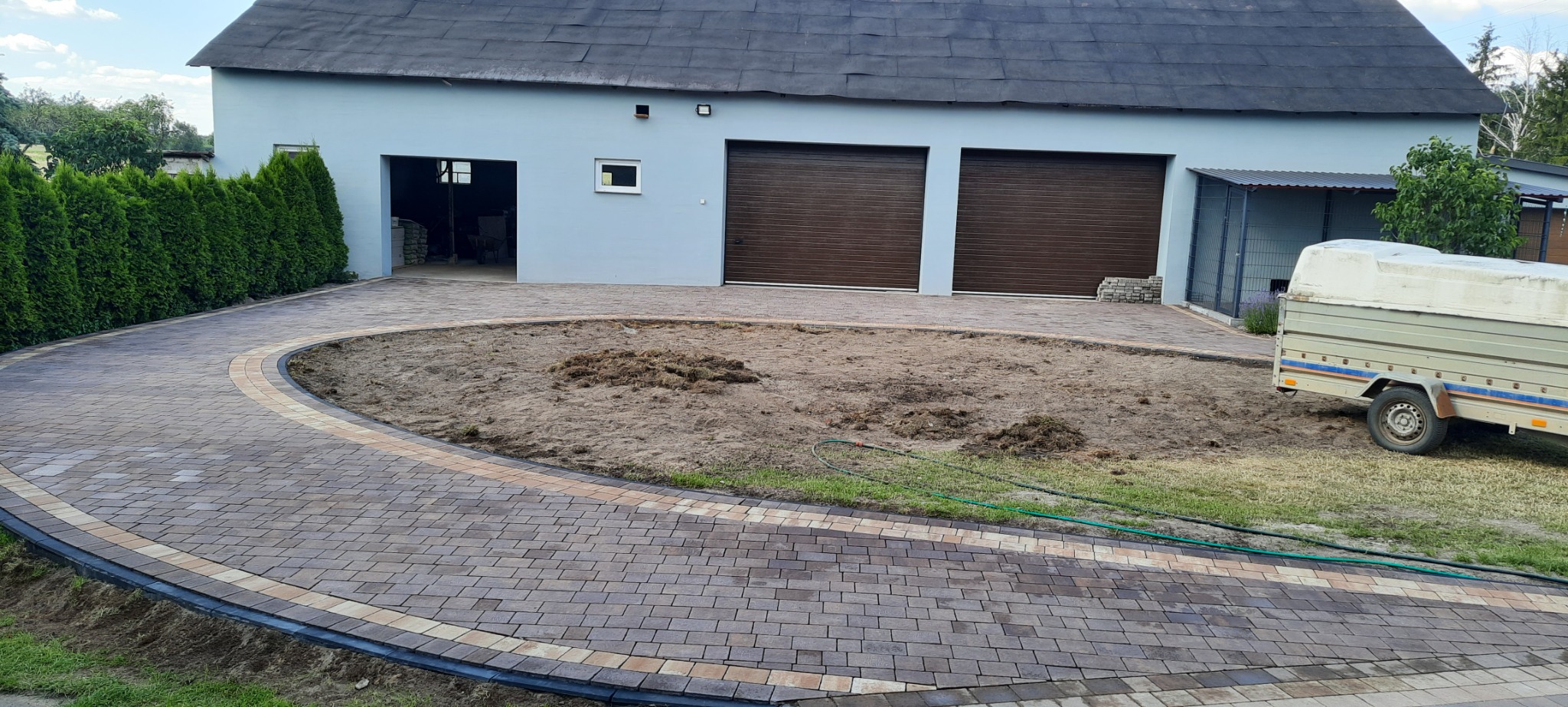 Ułożona, zakrzywiona ścieżka z kostki brukowej prowadząca do budynku z garażami, z centralnym placem w trakcie przygotowania pod trawnik lub nasadzenia, widoczny przyczepka.