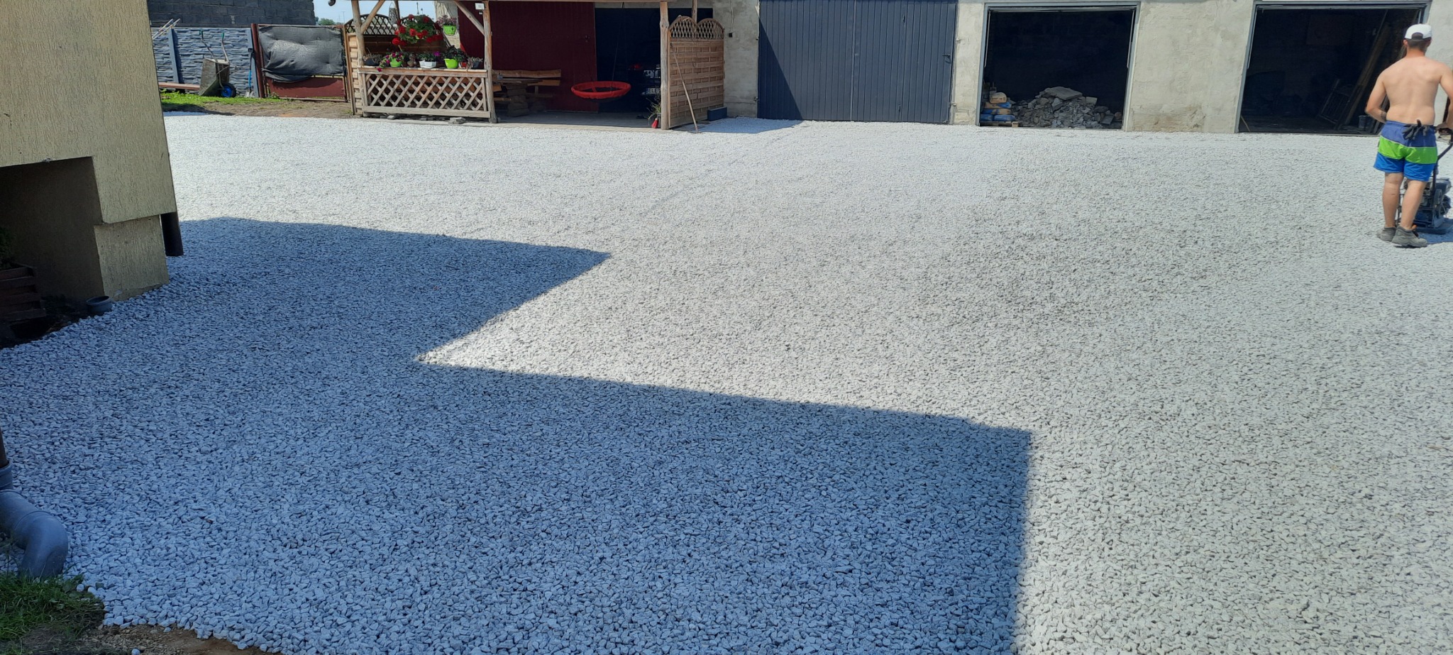 Utwardzona droga żwirowa z ubijarką w tle, widoczne budynki gospodarcze i fragment domu.