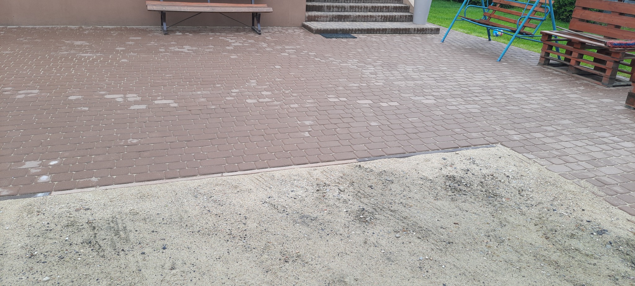 Fragment ułożonej, brązowej kostki brukowej z widocznym piaskiem do zasypywania szczelin, w tle ławka, schody i huśtawka.