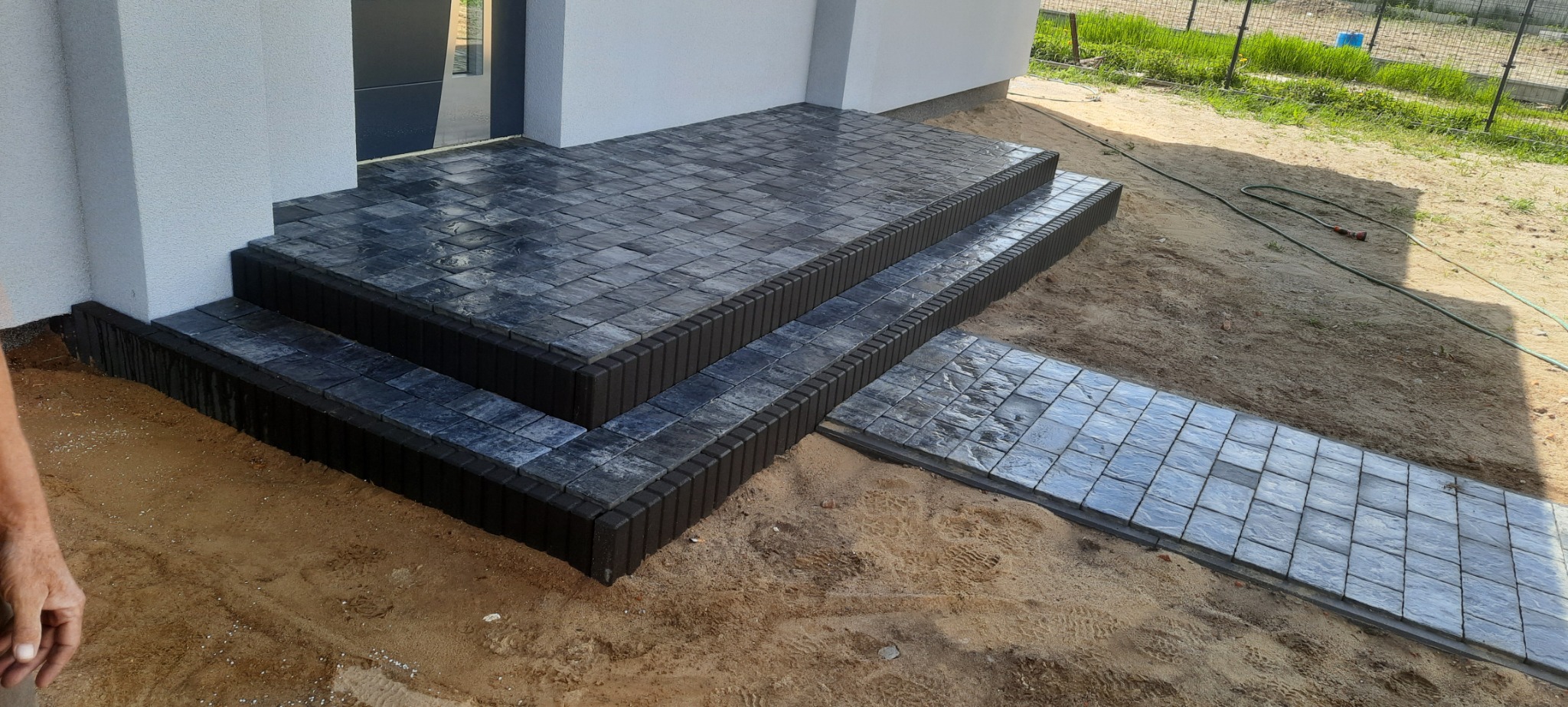 Schody wejściowe do domu wyłożone szarą kostką brukową z ciemnobrązowym obramowaniem, prowadzące do ścieżki z kostki, otoczone piaskiem i zielenią w tle.