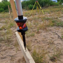 Biuro Geodezyjne mgr inż. Ernest Pilarski - Geodezyjny tycznik pryzmatyczny ustawiony na desce, oznaczający punkt tyczenia domu jednorodzinnego na piaszczystym terenie z trawą, w tle żółty teodolit na statywie.