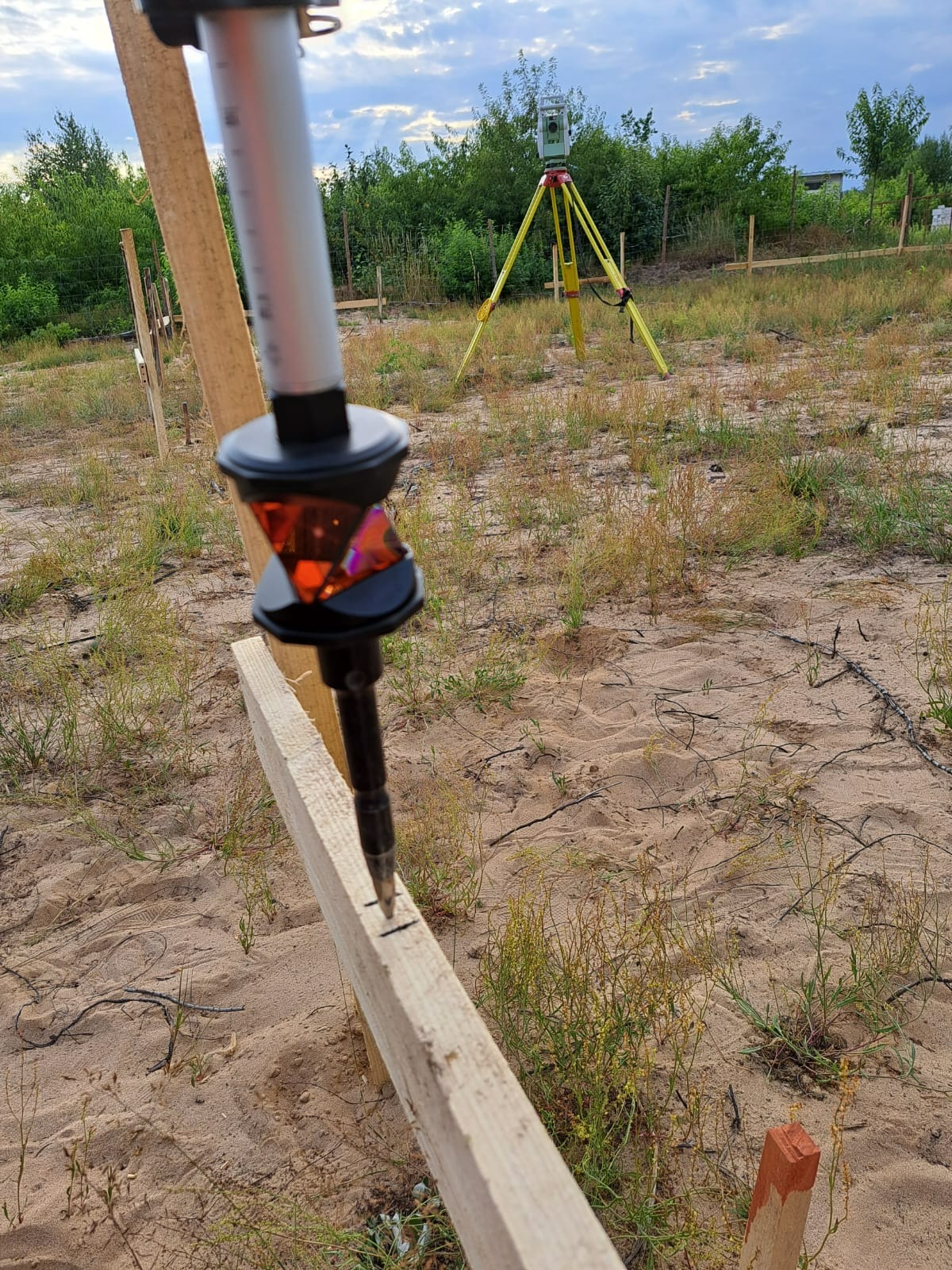 Geodezyjny tycznik pryzmatyczny ustawiony na desce, oznaczający punkt tyczenia domu jednorodzinnego na piaszczystym terenie z trawą, w tle żółty teodolit na statywie.