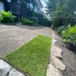 Fragment układania trawy z rolki w ogrodzie przydomowym, widoczny świeży fragment trawnika obok ścieżki z kamieni, w tle dom z drewnianą balustradą.