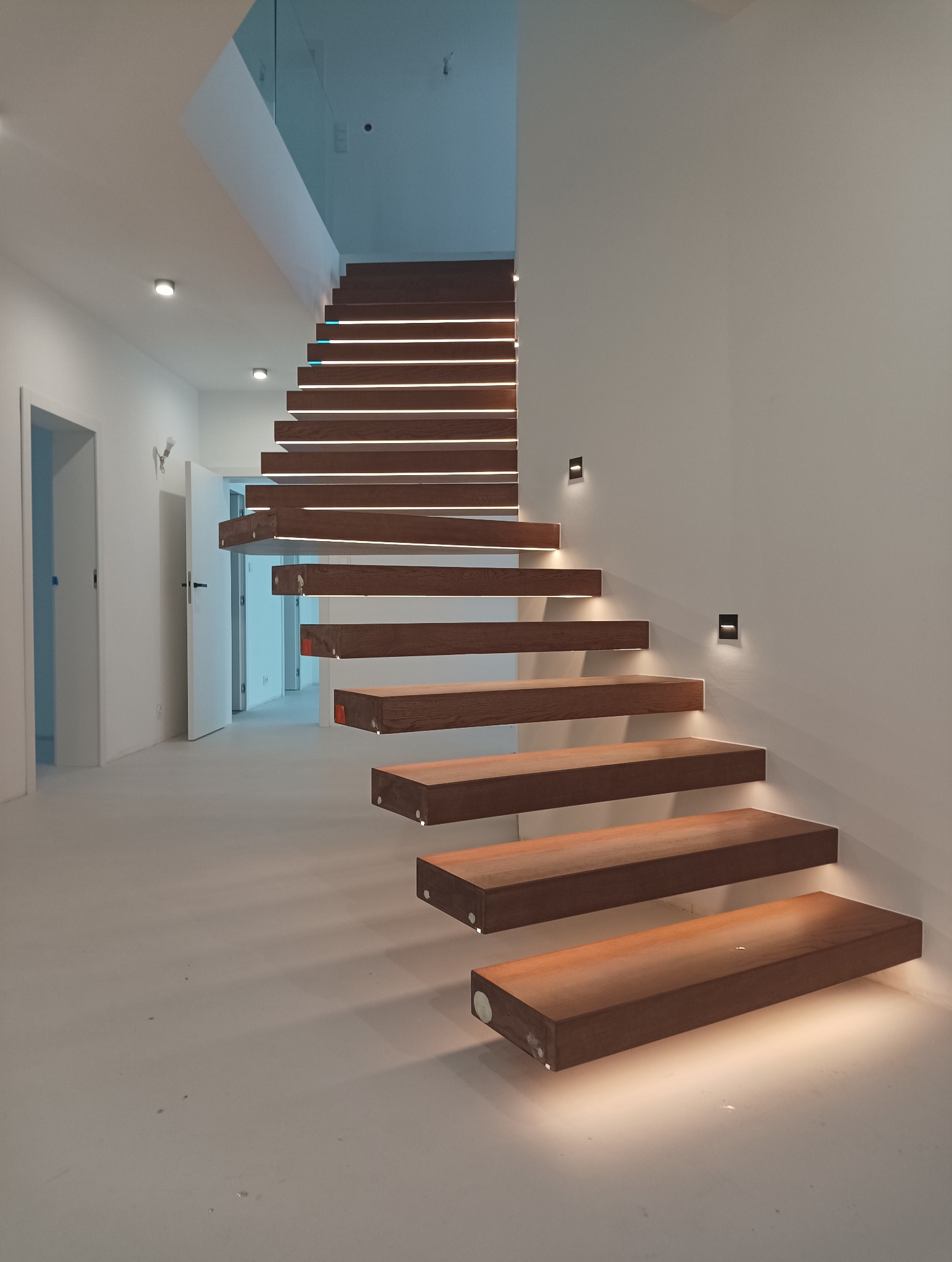 Nowoczesne schody drewniane z subtelnym oświetleniem LED pod każdym stopniem oraz punktowymi lampami ściennymi, minimalistyczny design w jasnym wnętrzu z przeszkloną balustradą.