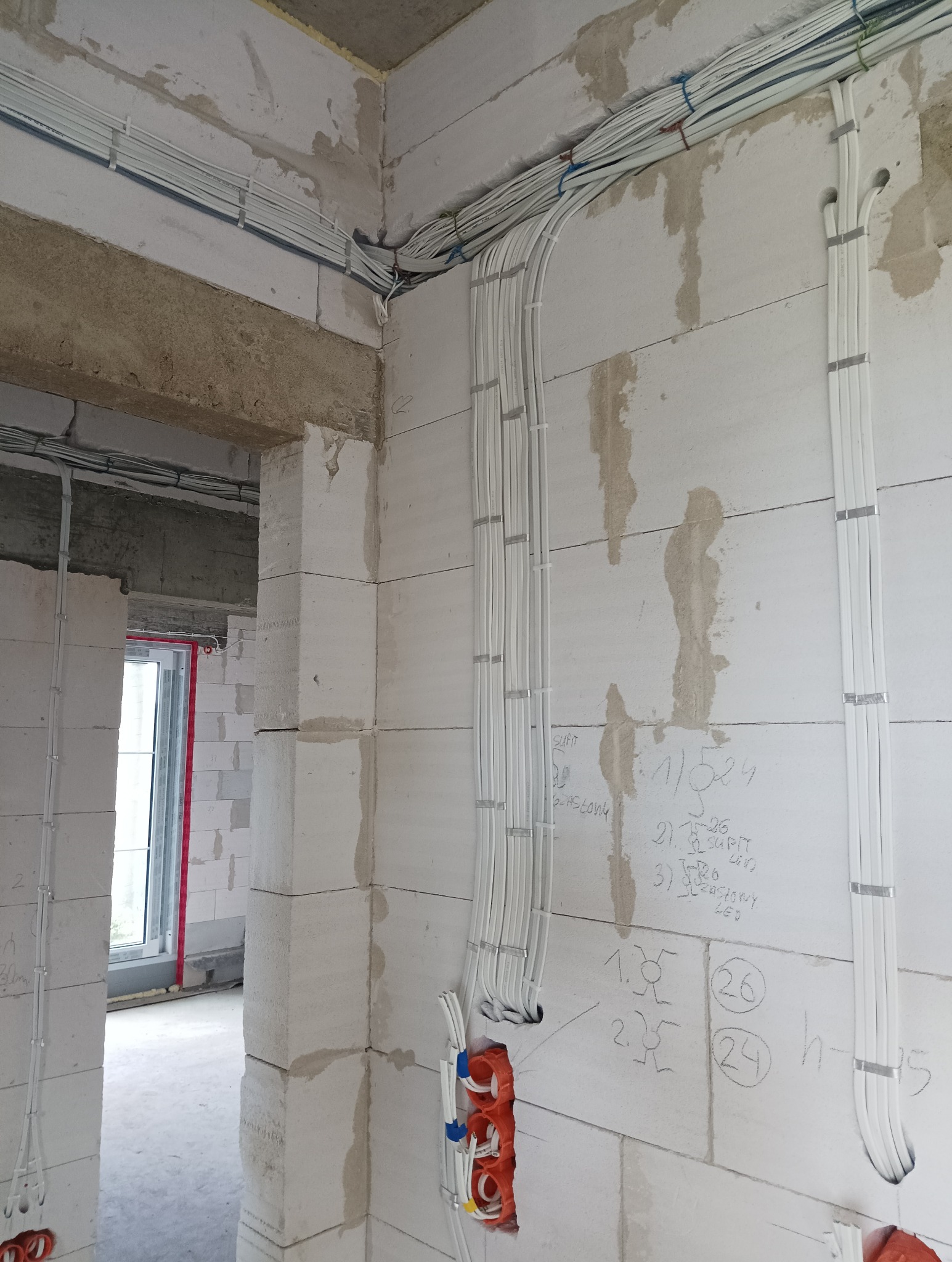 Nowa instalacja elektryczna w surowym domu jednorodzinnym, widoczne kable prowadzone w ścianach z betonu komórkowego, zabezpieczone uchwytami, puszki elektryczne z przewodami, na ścianie notatki...