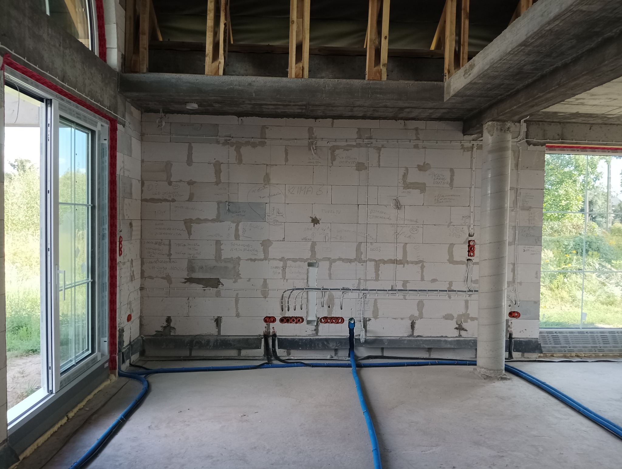 Wnętrze domu jednorodzinnego w trakcie budowy: widoczne instalacje elektryczne rozprowadzone po ścianach z bloczków, oznaczenia na ścianach, okno oraz niebieskie rury instalacyjne na podłodze.