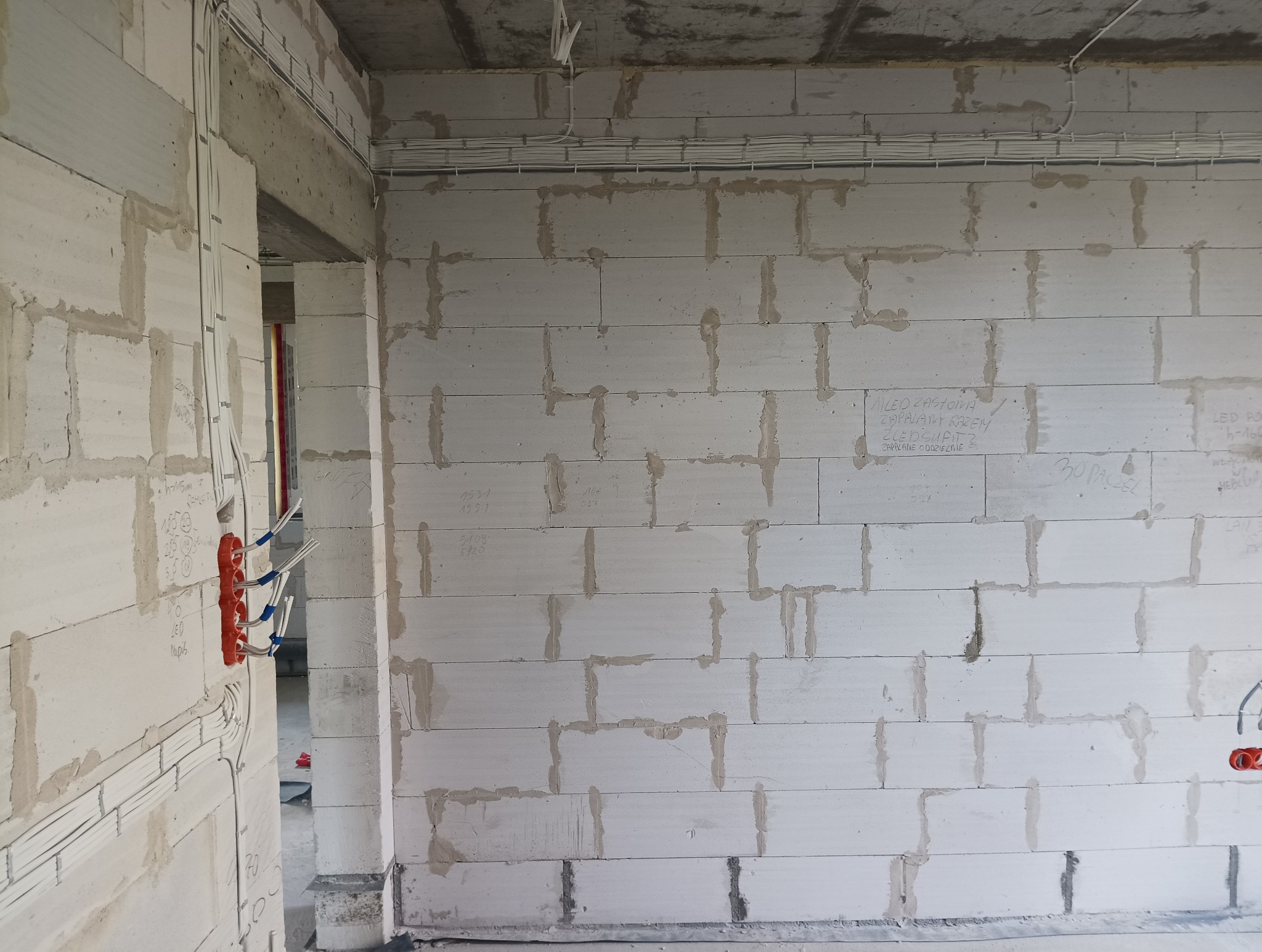 Instalacja elektryczna w nowym domu jednorodzinnym: widoczne przewody elektryczne prowadzone po ścianach z betonu komórkowego, puszki instalacyjne w kolorze pomarańczowym, oraz kable ułożone...