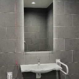 Łazienka z szarymi płytkami, umywalką z uchwytami dla niepełnosprawnych i podświetlanym lustrem.