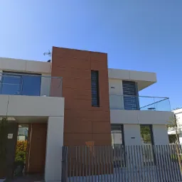 Nowoczesny dom z białymi ścianami, drewnianą elewacją i szklanymi balustradami balkonowymi, widziany z zewnątrz za metalowym ogrodzeniem.