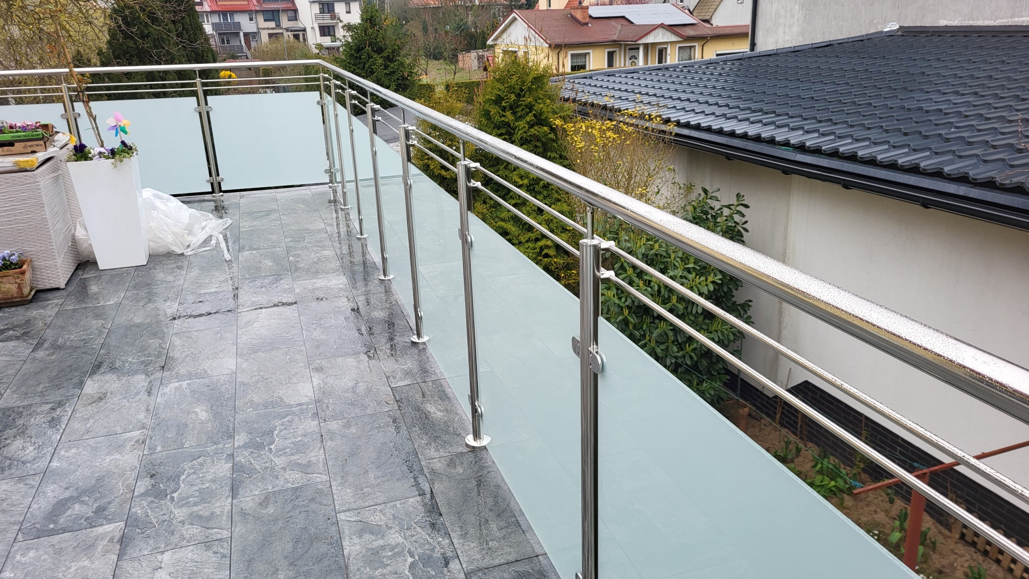 Balkon z szarymi płytkami i nowoczesną balustradą ze stali nierdzewnej i szkła matowego, widok na ogród i domy w tle.