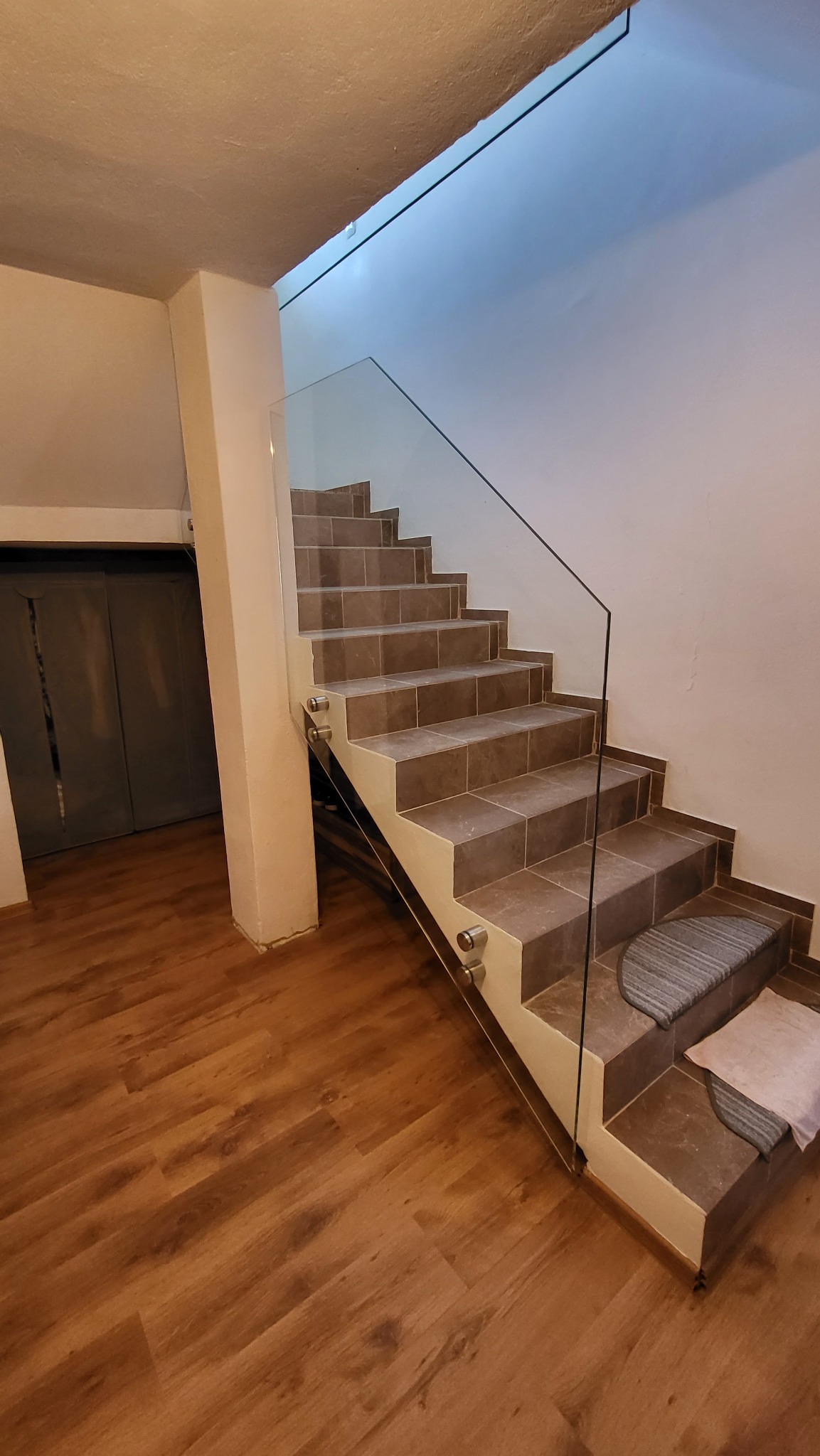 Schody wyłożone brązowymi płytkami z przezroczystą szklaną balustradą mocowaną na metalowych uchwytach, widok z dołu na drewnianą podłogę.