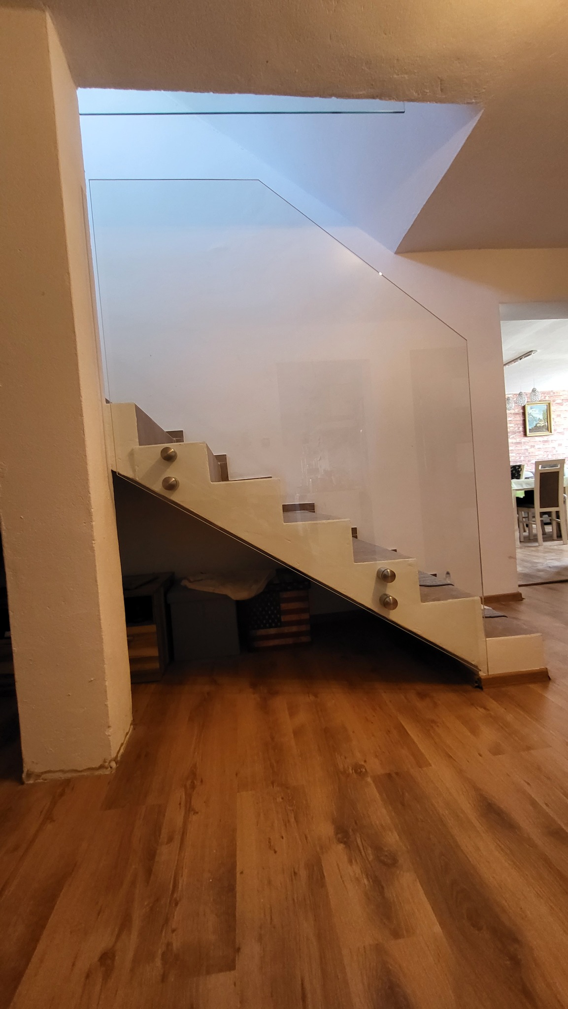 Szklana balustrada zamocowana do schodów za pomocą metalowych uchwytów, widok z dołu na drewnianą podłogę i fragment kolumny.