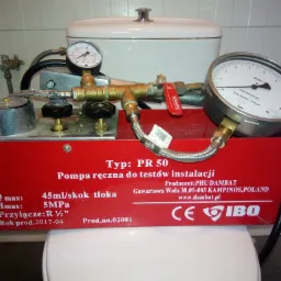 Czerwona pompa ręczna PR 50 do testów instalacji, producent PHU Dambat, z dwoma manometrami, podłączona do instalacji wodnej, ustawiona na sedesie w łazience.