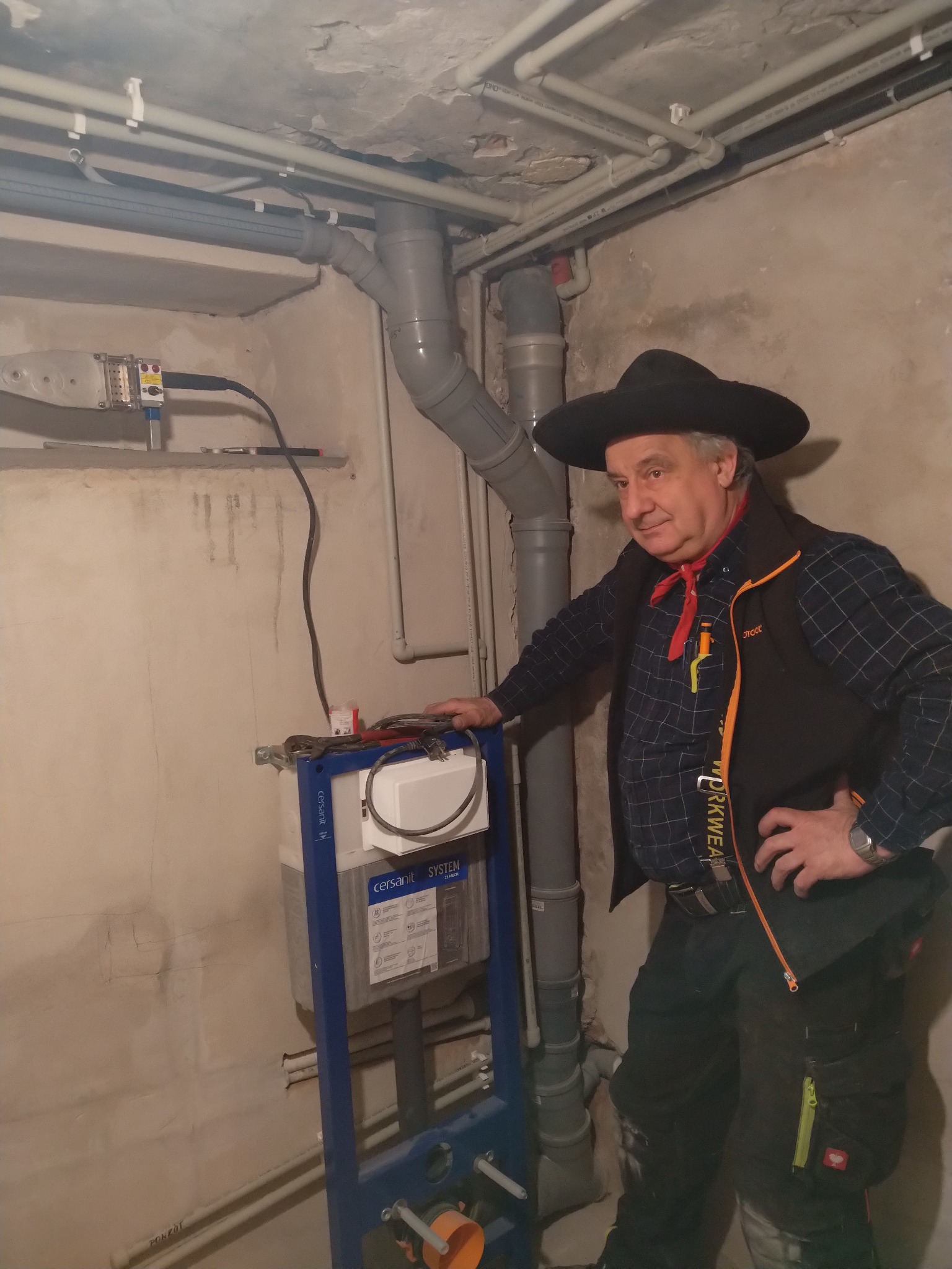 Hydraulik w kapeluszu kowbojskim prezentuje zamontowany stelaż podtynkowy WC Cersanit System, widoczne rury kanalizacyjne i instalacje wodne w surowym wnętrzu.