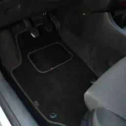 Czarne, welurowe dywaniki samochodowe z czerwono-białym obszyciem, leżące na podłodze auta, widoczne pedały i fragment fotela.