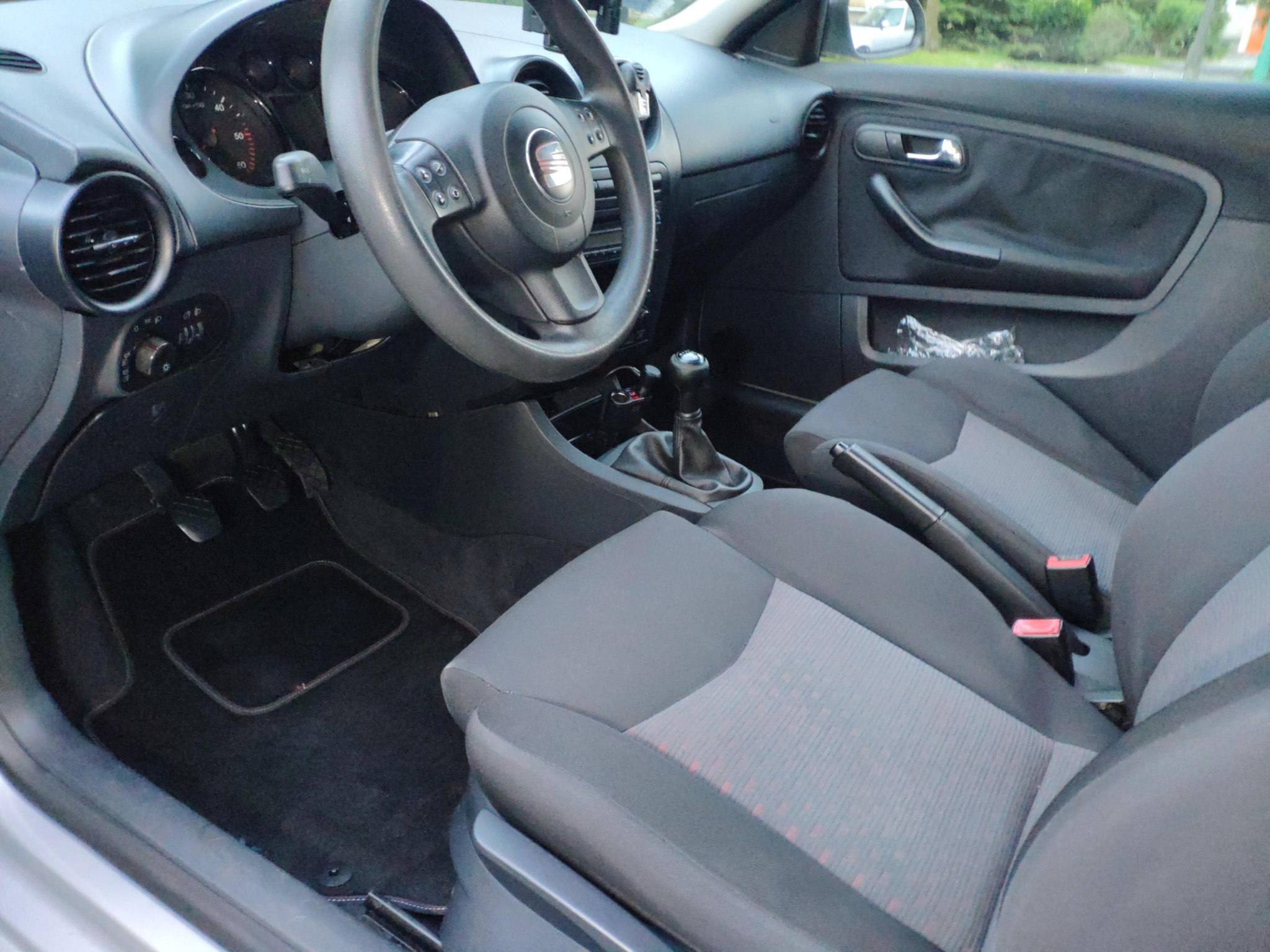 Wnętrze samochodu marki Seat Ibiza z ciemną tapicerką, widoczne fotele z szaro-czarnym wzorem, kierownica z logo, deska rozdzielcza i czarne dywaniki.