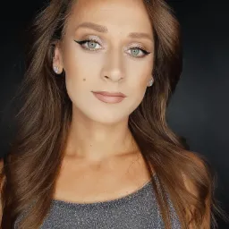 Portret kobiety z delikatnym, błyszczącym cieniem na powiekach i precyzyjną kreską eyelinerem, uzupełnionym o subtelny odcień szminki i srebrzystą biżuterię na tle ciemnego tła.