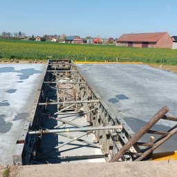Marona Sebastian Usługi Ogólnobudowlane - Wylany fundament z szalunkiem i drewnianą drabiną opartą o krawędź. W tle pola i zabudowania wiejskie w słoneczny dzień.