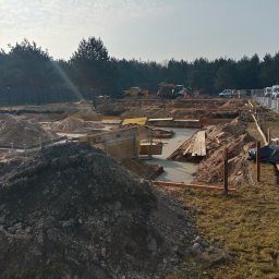 Marona Sebastian Usługi Ogólnobudowlane - Wykop pod fundamenty domu w trakcie prac ziemnych. Widoczne szalunki, wykopane rowy, hałdy ziemi, sprzęt budowlany w tle i ogrodzenie terenu budowy.