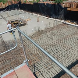Marona Sebastian Usługi Ogólnobudowlane - Zalewanie betonem zbrojonej płyty fundamentowej. Widoczny pracownik w żółtych butach i stalowe szalunki. Ujęcie z góry, z rusztowania.