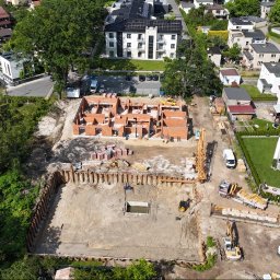 Marona Sebastian Usługi Ogólnobudowlane - Zdjęcie z lotu ptaka przedstawia plac budowy z częściowo wzniesionymi ścianami z czerwonej cegły, zabezpieczonym wykopem i ciężkim sprzętem budowlanym. W tle zabudowa mieszkalna.