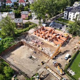Marona Sebastian Usługi Ogólnobudowlane - Widok z lotu ptaka na plac budowy z wykopem zabezpieczonym ściankami Larsena i wznoszoną konstrukcją domu z czerwonej cegły. Na placu budowy widoczne maszyny budowlane.